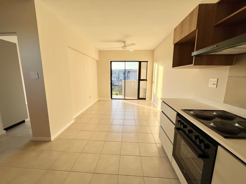 An open-plan living and kitchen area featuring tiled floors, built-in cabinetry with an integrated oven and hob, and access to a balcony via sliding doors.