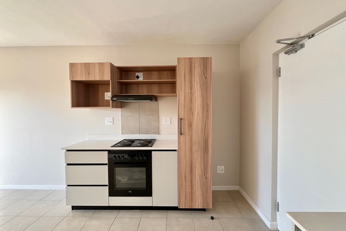 A modern, compact kitchen featuring light wood-tone cabinetry, a built-in oven, electric hob, extractor fan, and neutral tiled flooring in an open-plan layout.