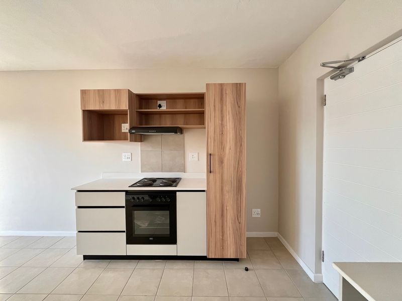 A modern, compact kitchen featuring light wood-tone cabinetry, a built-in oven, electric hob, extractor fan, and neutral tiled flooring in an open-plan layout.