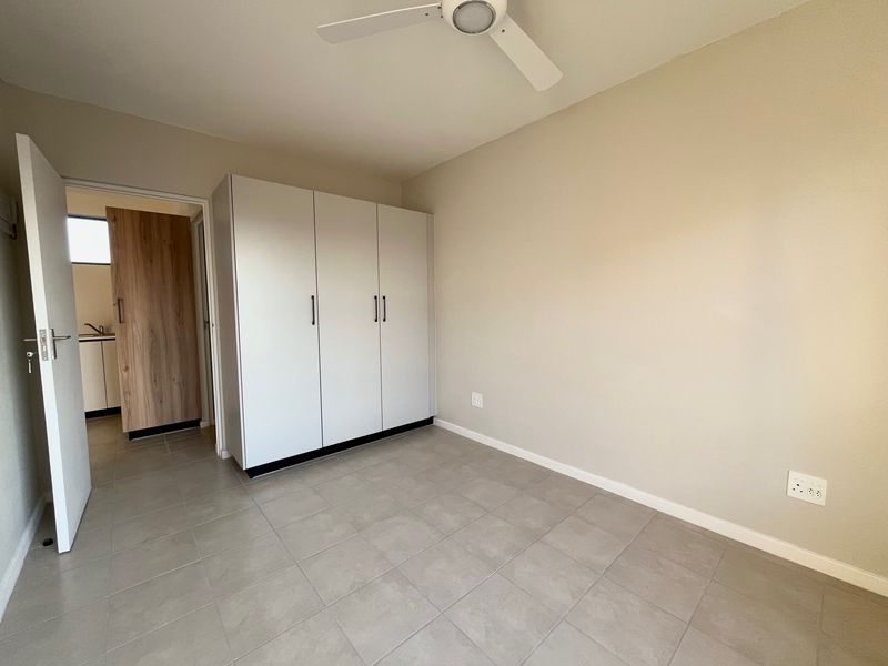 A clean, empty bedroom featuring neutral tiled flooring, built-in white cupboards, and a ceiling fan, with a view into an adjacent room.