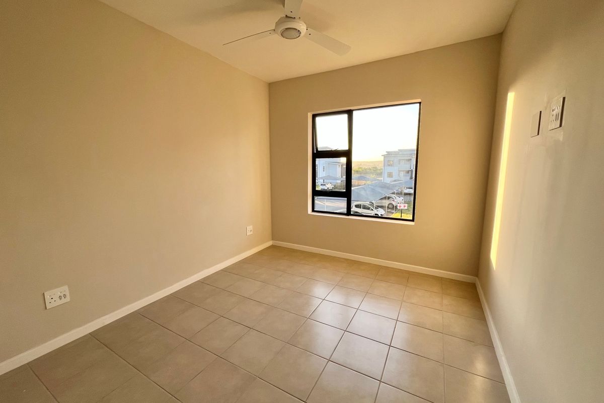 A clean, empty bedroom featuring neutral tiled flooring, a ceiling fan, and a large window providing natural light and a view of the complex.