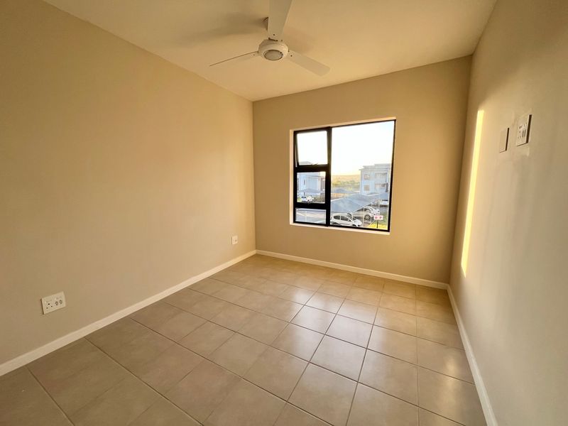 A clean, empty bedroom featuring neutral tiled flooring, a ceiling fan, and a large window providing natural light and a view of the complex.
