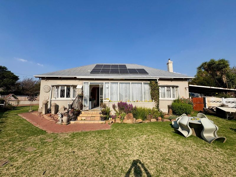 Rear view of a single-story house featuring a corrugated metal roof with installed solar panels, bay windows, and a well-maintained garden area.
