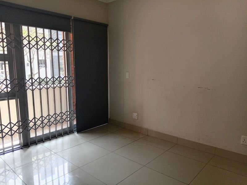 An empty living area featuring tiled flooring, a sliding glass door with a security gate, and roller blinds, ready for personalization.