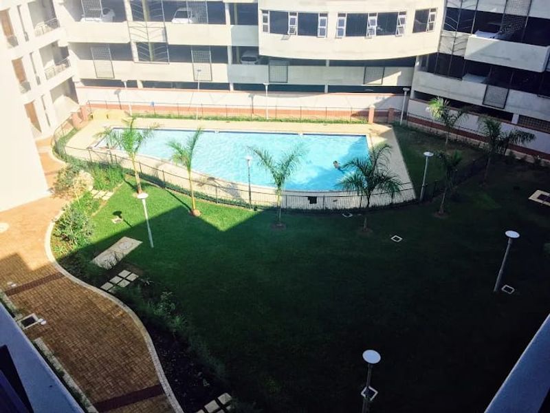 An elevated view of a communal swimming pool area featuring a landscaped lawn, paved pathways, and perimeter fencing within an apartment complex.