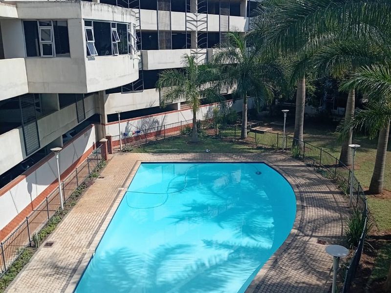 An elevated view of a communal swimming pool area featuring a paved deck, perimeter fencing, and established palm trees within a residential complex.