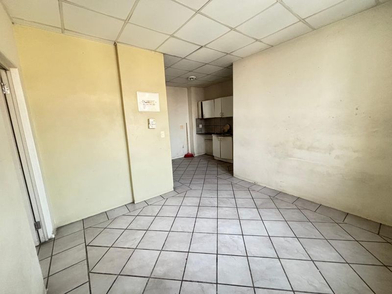 An empty open-plan living and kitchen area featuring tiled flooring and a drop ceiling, requiring cosmetic updates and wall repairs.