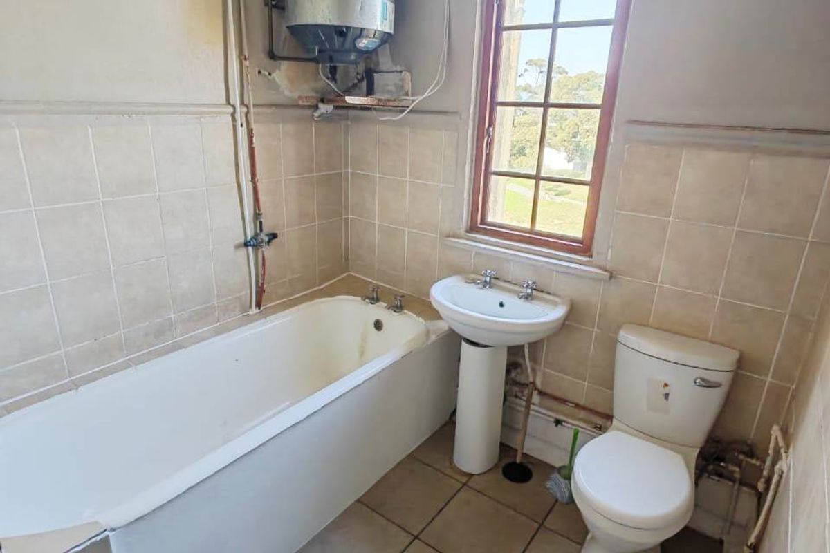 A functional bathroom featuring a bathtub, pedestal basin, and toilet, with an exposed electric water heater and dated ceramic tiling throughout.