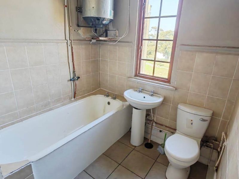 A functional bathroom featuring a bathtub, pedestal basin, and toilet, with an exposed electric water heater and dated ceramic tiling throughout.