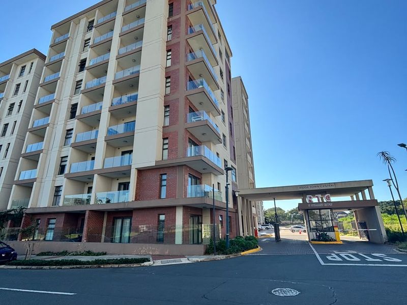 Exterior view of a modern multi-story apartment complex featuring glass balconies, brick detailing, and a secure gated entrance.