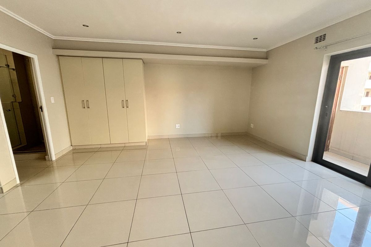 Spacious, empty bedroom featuring neutral tiled flooring, built-in cupboards, recessed lighting, and a sliding glass door providing access to a balcony.