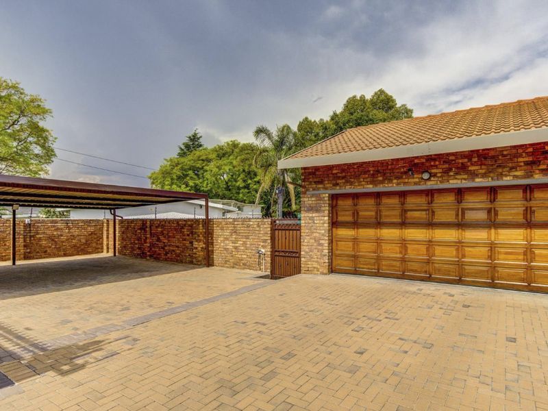 Front exterior view featuring a paved driveway, a double garage with wooden doors, and a covered carport area with brick boundary walls.