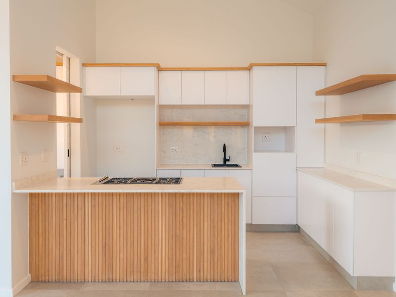 Modern kitchen featuring white cabinetry, timber slat island, stone countertops, floating shelves, and integrated gas hob in a bright, open-plan space.