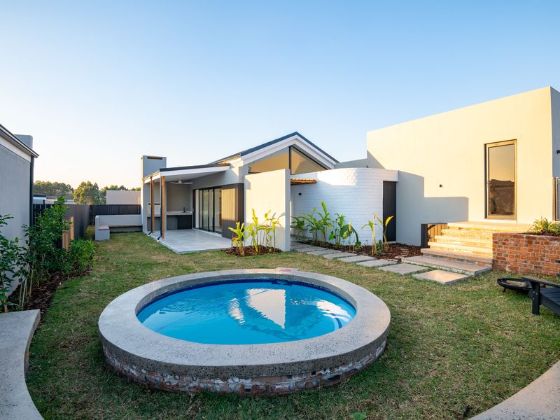 A modern backyard featuring a circular plunge pool, a covered patio with a built-in braai, and landscaped garden areas.