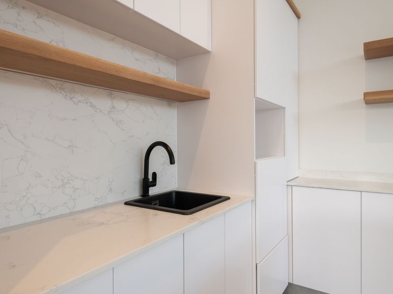 Modern scullery featuring white cabinetry, marble-look quartz countertops, a black sink with matching faucet, and timber floating shelves.