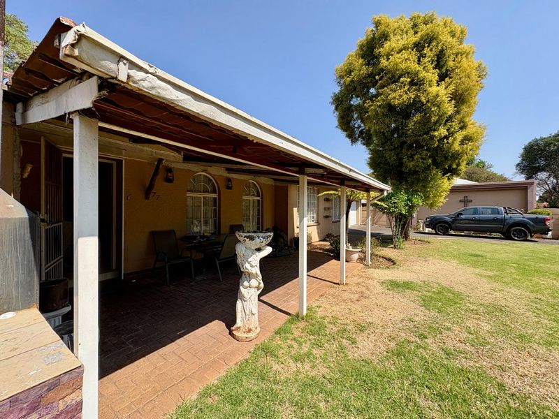A covered brick-paved patio area featuring arched windows and a timber roof structure, overlooking a spacious garden.