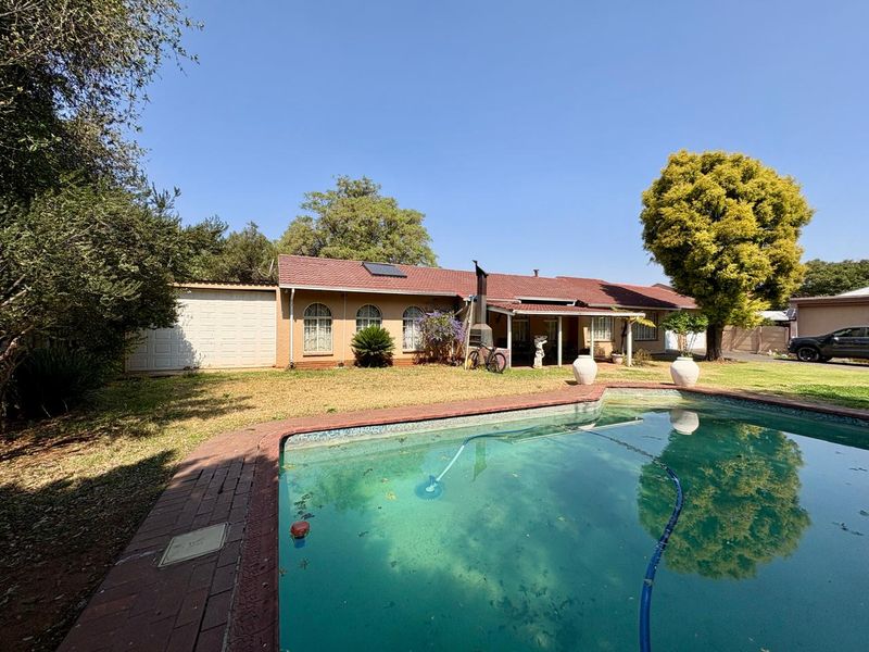 Rear exterior view of the house featuring a swimming pool, covered patio area, and established garden surroundings.