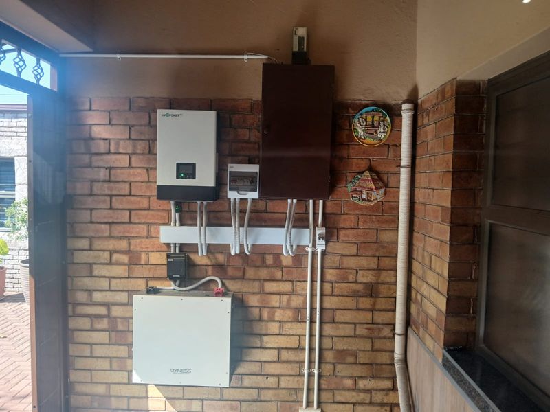 Utility area featuring a wall-mounted solar inverter and battery storage system with organized electrical conduit and distribution board on an exposed brick wall.