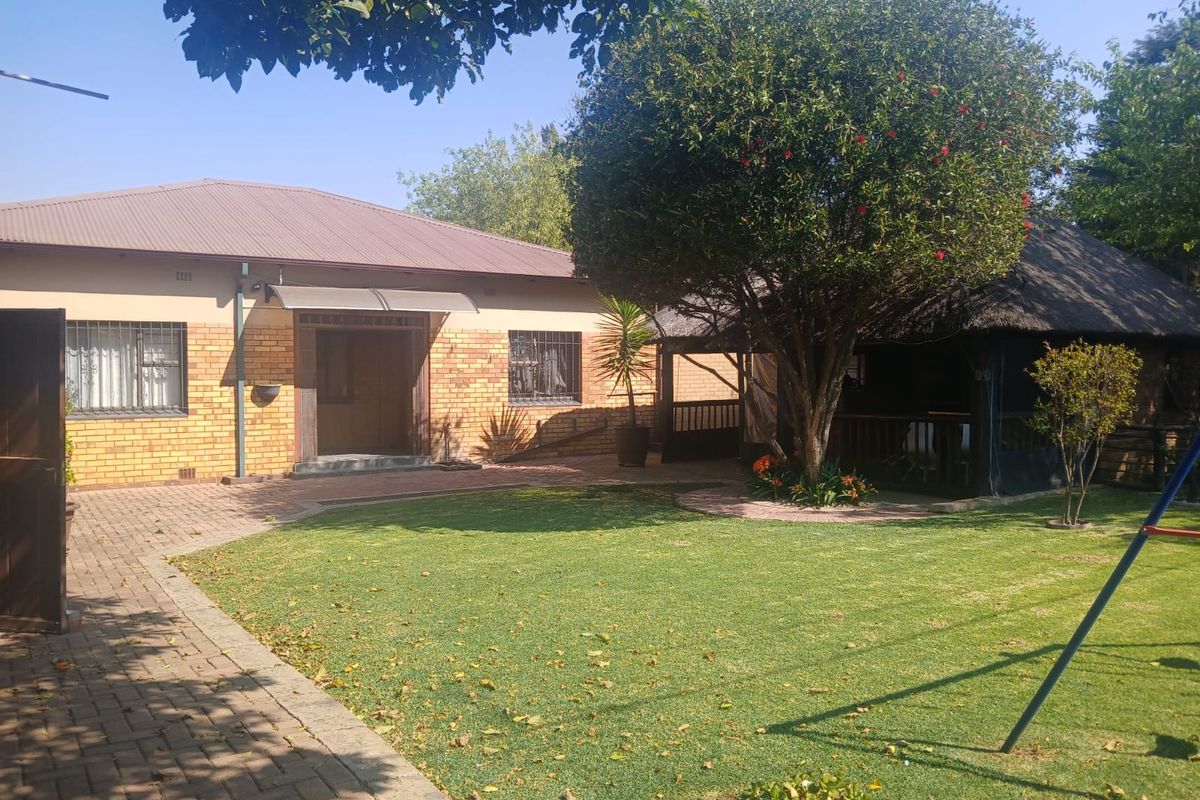 Rear view of the property featuring a brick facade, metal roof, paved area, and a thatched lapa set within a well-maintained garden.