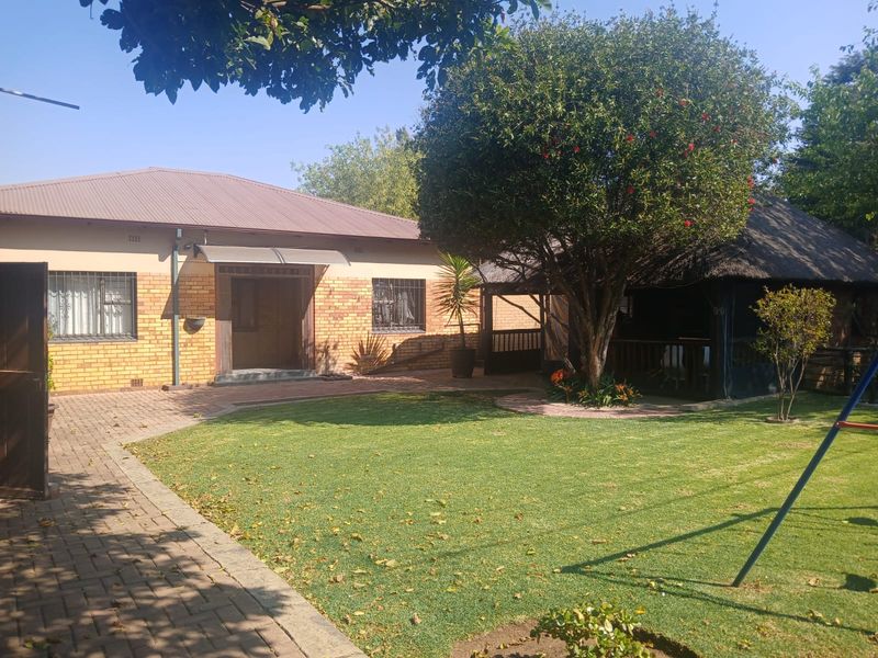 Rear view of the property featuring a brick facade, metal roof, paved area, and a thatched lapa set within a well-maintained garden.