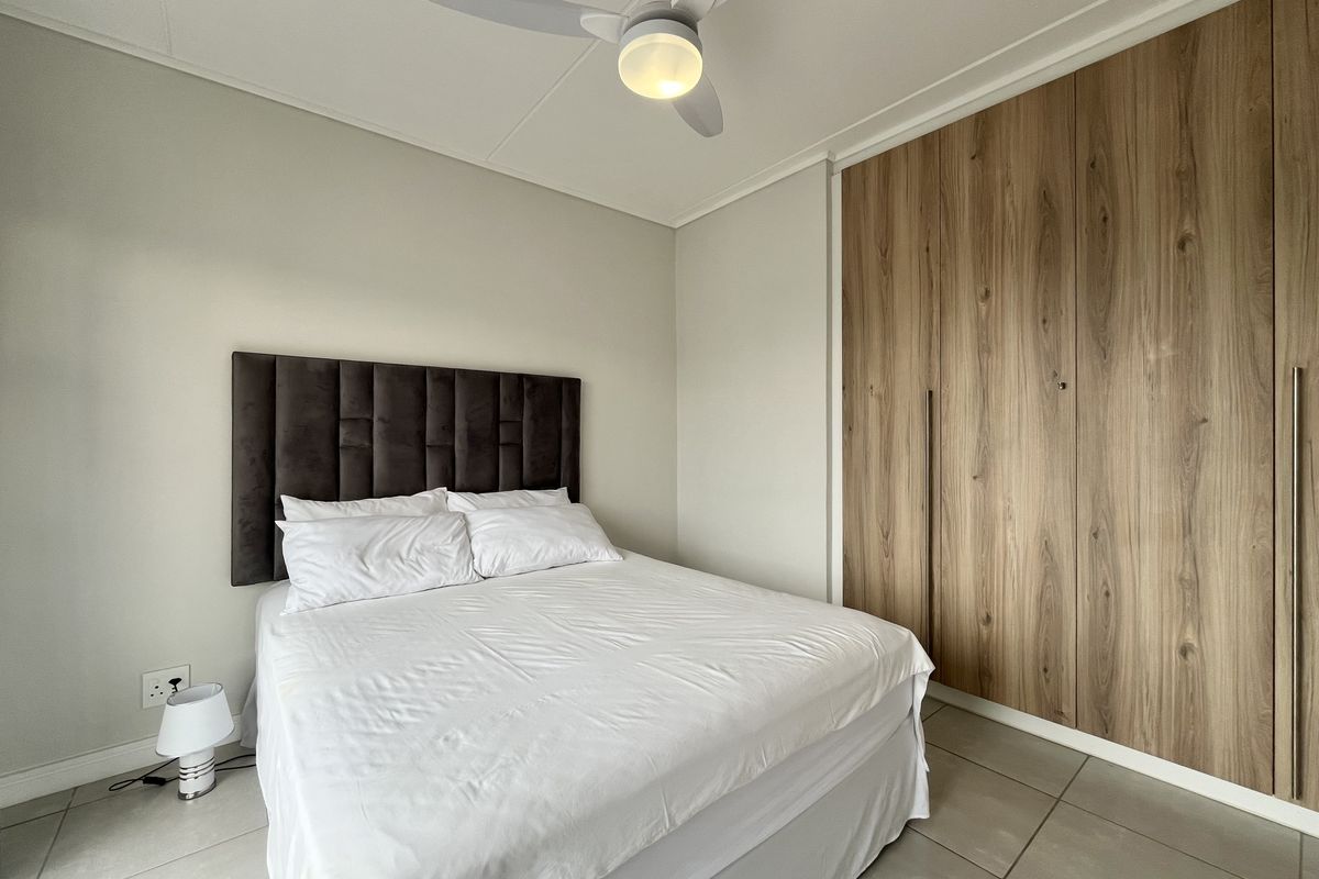 A well-maintained bedroom featuring modern wood-finish built-in cupboards, a ceiling fan, and neutral tiled flooring.