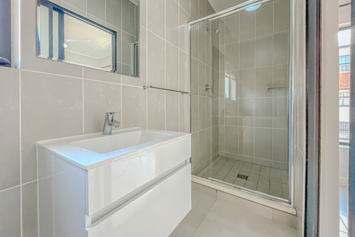 A clean, modern bathroom featuring a white vanity cabinet, basin, wall-mounted mirror, and a glass-enclosed shower with neutral wall and floor tiling.