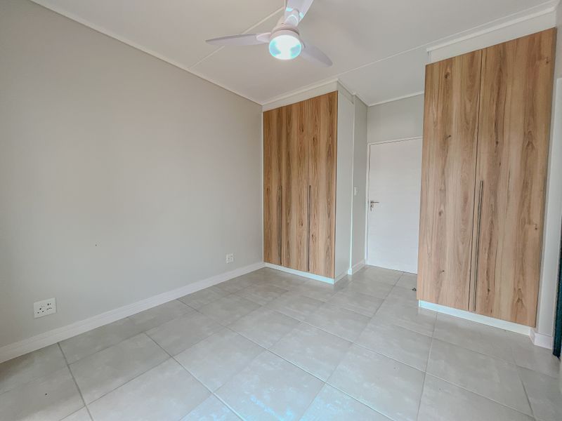 A bright, empty bedroom featuring modern wood-finish built-in cupboards, neutral tiled flooring, and a ceiling fan.