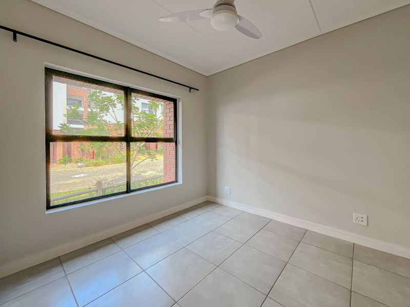 A bright, empty bedroom featuring neutral tiled flooring, a ceiling fan, and a large window providing natural light and a view of the complex grounds.