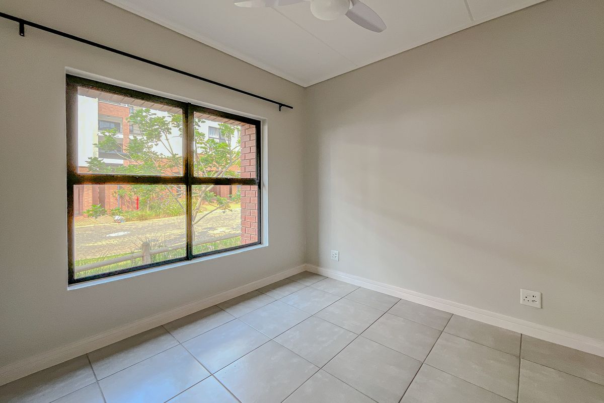 A bright, empty bedroom featuring neutral tiled flooring, a ceiling fan, and a large window providing natural light and a view of the complex grounds.