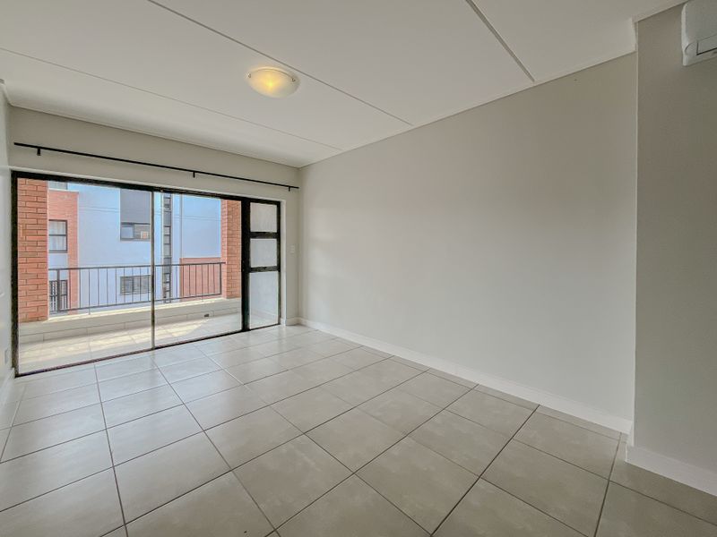 A bright, empty living room featuring neutral tiled flooring, a wall-mounted air conditioning unit, and a large sliding glass door leading to a balcony.