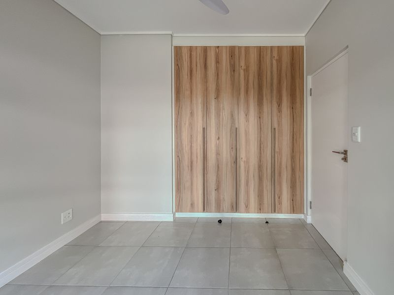 A clean, empty bedroom featuring modern wood-finish built-in cupboards, neutral grey tiled flooring, and a ceiling fan.
