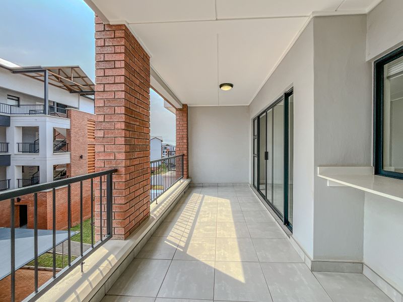 A spacious covered balcony featuring tiled flooring, exposed brick columns, and a metal balustrade, providing a clean and functional outdoor living space.