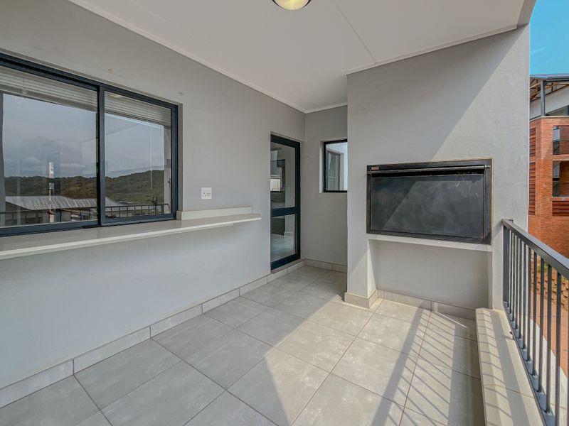 A clean, covered balcony featuring tiled flooring, a built-in braai, and a metal balustrade, offering a functional outdoor space for relaxation.