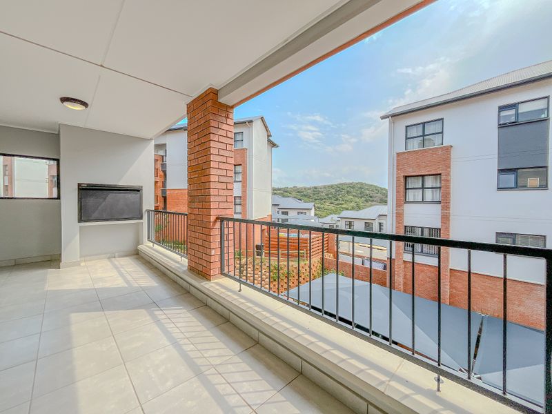 A spacious covered balcony featuring tiled flooring, a built-in braai, and a metal balustrade, offering pleasant views of the surrounding apartment complex.