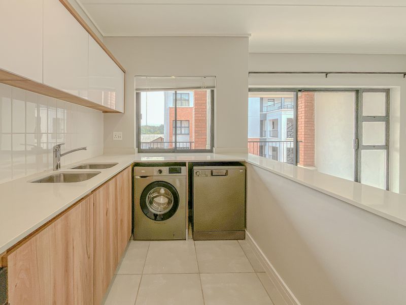 A clean and modern scullery area featuring light wood cabinetry, stone countertops, a stainless steel sink, and space for under-counter appliances.