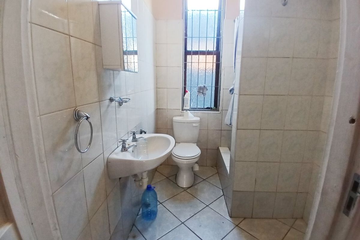 A functional bathroom featuring a wall-mounted basin, toilet, and shower enclosure with tiled finishes and a window with security bars.