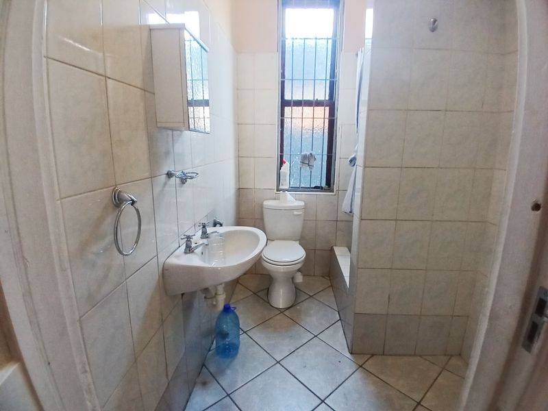 A functional bathroom featuring a wall-mounted basin, toilet, and shower enclosure with tiled finishes and a window with security bars.