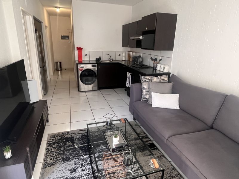 An open-plan living area featuring a compact kitchen with dark cabinetry, integrated appliances, and tiled flooring, seamlessly connected to the lounge space.