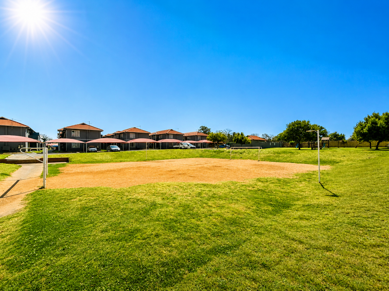 A communal sports field and recreational area featuring a grass pitch and adjacent shade cloth parking structures within the apartment complex.