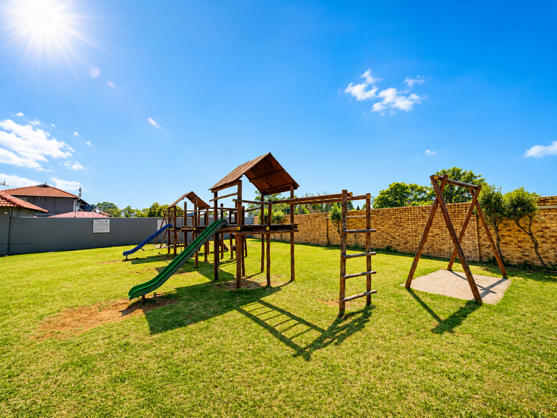 A well-maintained communal garden area featuring a wooden playground structure with slides and swings, enclosed by a brick boundary wall.