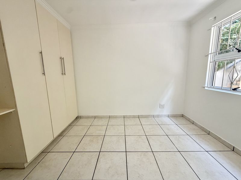 A bright, empty bedroom featuring neutral tiled flooring, built-in cupboards, and a window fitted with security bars.