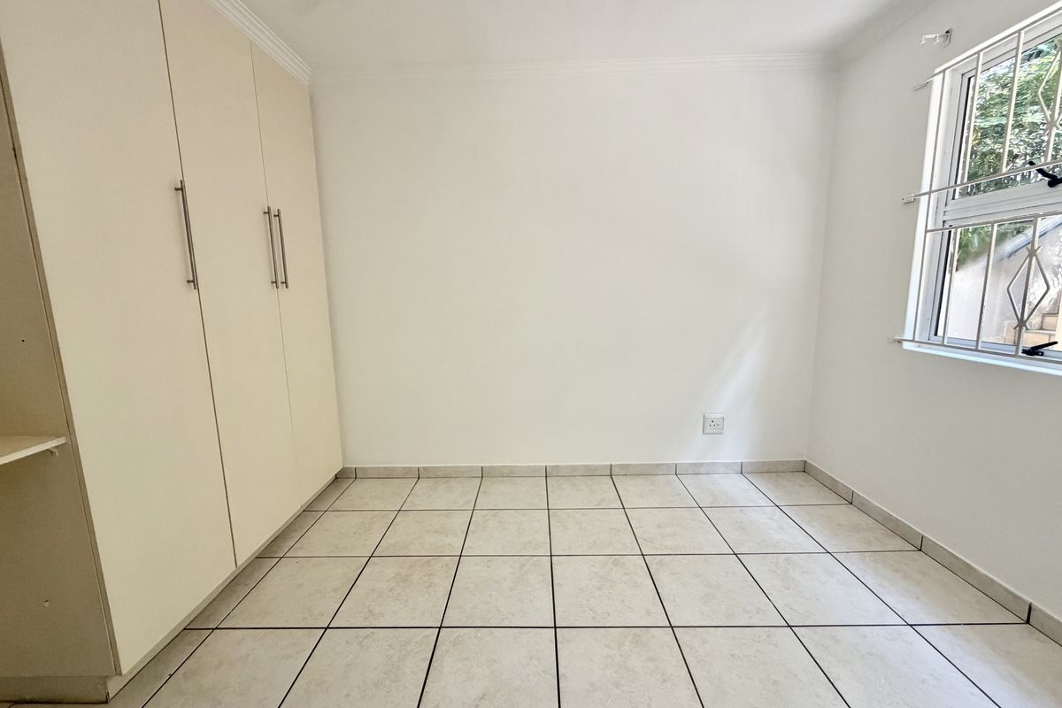A bright, empty bedroom featuring neutral tiled flooring, built-in cupboards, and a window fitted with security bars.
