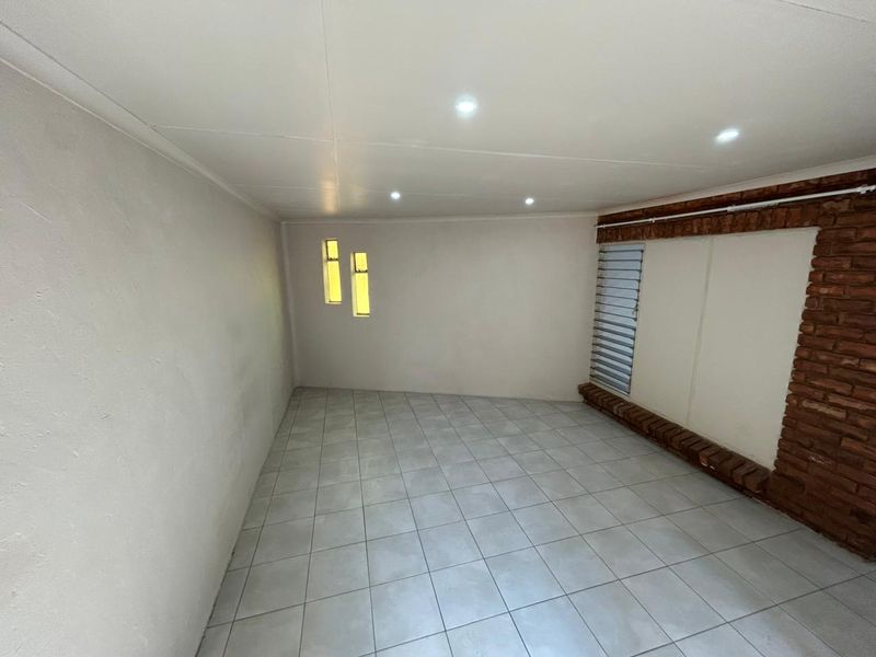 An empty living area featuring tiled flooring, recessed downlights, and an exposed brick feature wall with louvered windows.