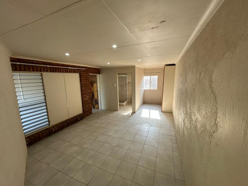 An empty bedroom featuring tiled floors, an exposed brick accent wall, recessed lighting, and an attached en-suite bathroom with a shower.
