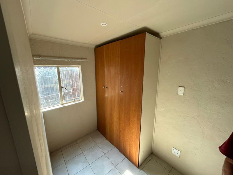 A compact bedroom featuring tiled flooring, a built-in wooden wardrobe, and a window fitted with security bars.
