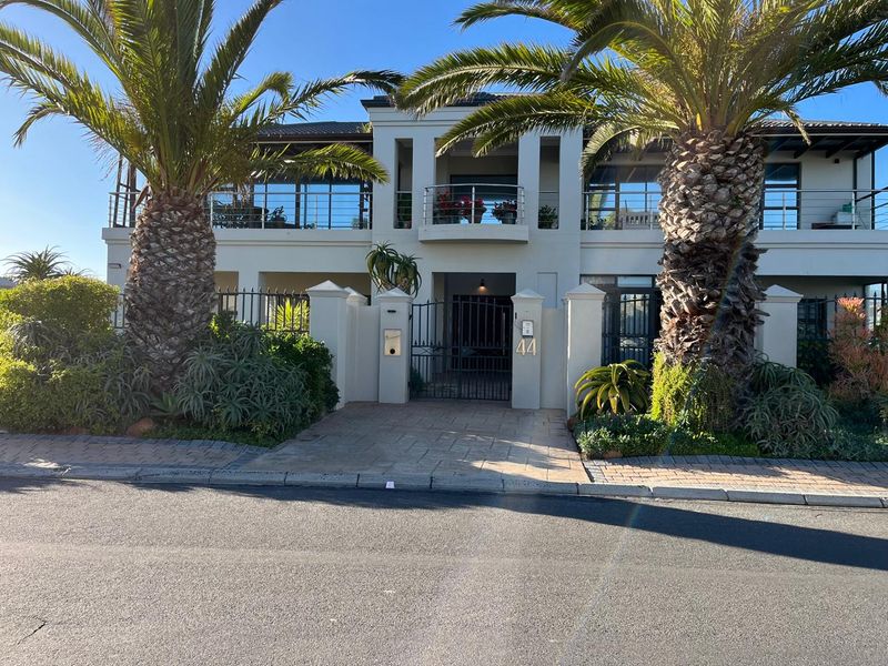 Front elevation of a two-story apartment building featuring a gated entrance, paved driveway, and upper-level balcony with stainless steel balustrades.