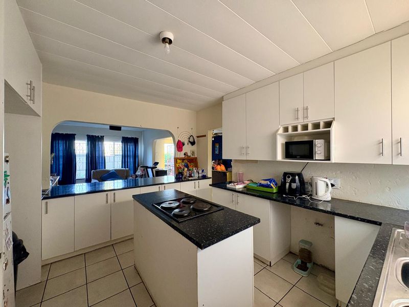 Functional kitchen featuring white cabinetry, dark granite countertops, a central island with an electric hob, and tiled flooring, opening into the living area.