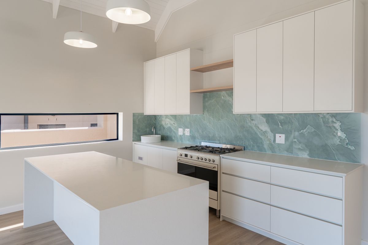 Modern kitchen featuring white cabinetry, stone countertops, a striking green stone slab backsplash, gas stove, and a central island under a vaulted ceiling.