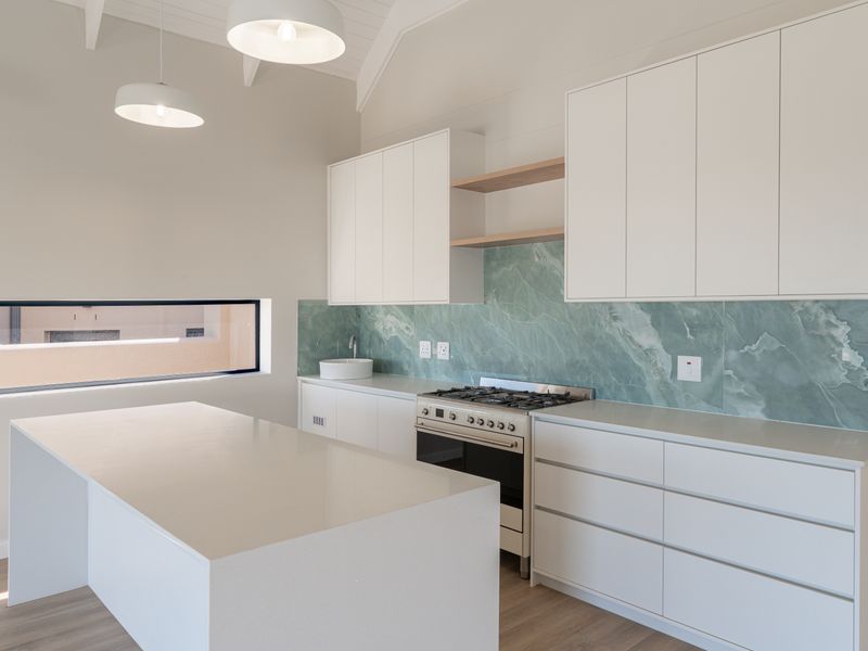 Modern kitchen featuring white cabinetry, stone countertops, a striking green stone slab backsplash, gas stove, and a central island under a vaulted ceiling.