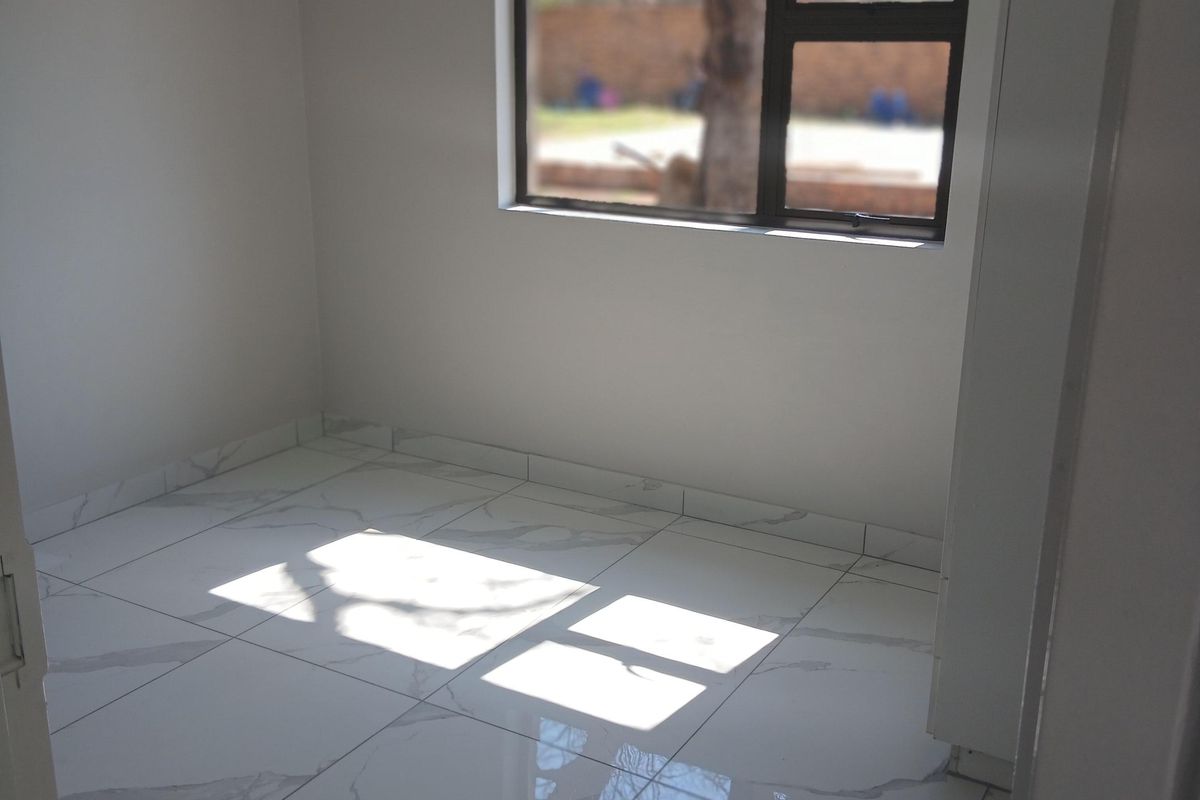 A bright, empty bedroom featuring modern white marble-look floor tiles and a dark aluminium window frame.
